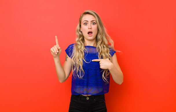 young blonde woman feeling proud and surprised, pointing to self confidently, feeling like successful number one against red wall - Фото, зображення