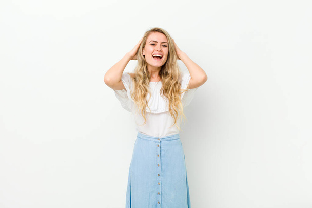 young blonde woman looking happy, carefree, friendly and relaxed enjoying life and success, with a positive attitude against white wall - Фото, изображение