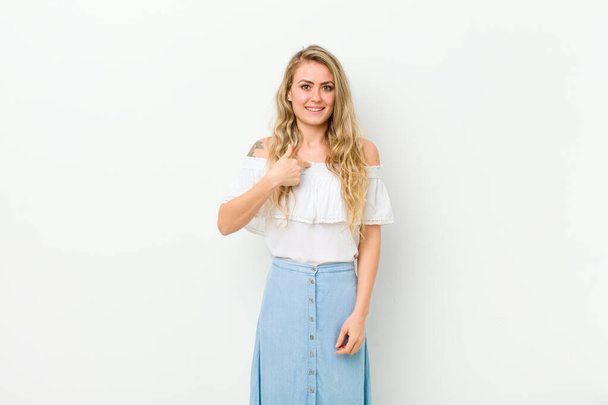 young blonde woman looking happy, proud and surprised, cheerfully pointing to self, feeling confident and lofty against white wall - Foto, afbeelding