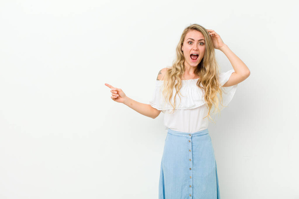 young blonde woman laughing, looking happy, positive and surprised, realizing a great idea pointing to lateral copy space against white wall - Photo, Image