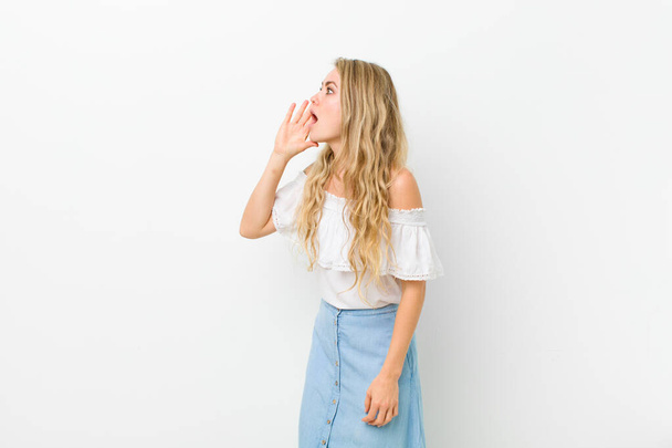 young blonde woman yelling loudly and angrily to copy space on the side, with hand next to mouth against white wall - Φωτογραφία, εικόνα