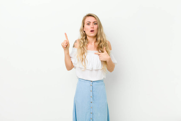 young blonde woman feeling proud and surprised, pointing to self confidently, feeling like successful number one against white wall - Fotografie, Obrázek