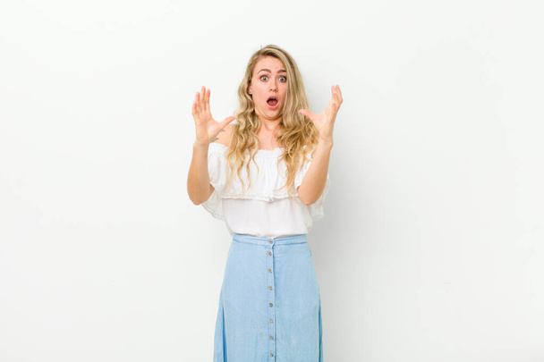 young blonde woman screaming with hands up in the air, feeling furious, frustrated, stressed and upset against white wall - Foto, imagen