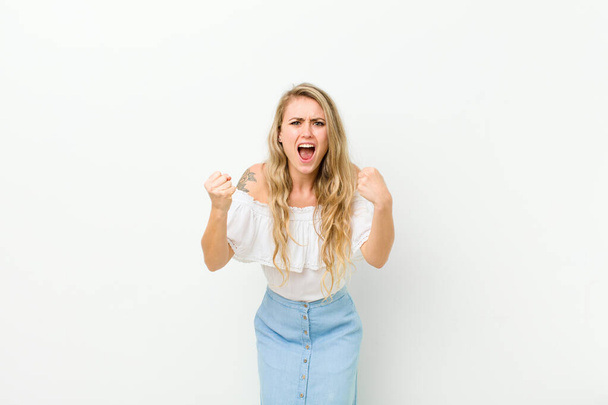 young blonde woman shouting aggressively with an angry expression or with fists clenched celebrating success against white wall - Fotografie, Obrázek
