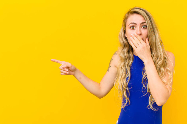 young blonde woman feeling happy, shocked and surprised, covering mouth with hand and pointing to lateral copy space against yellow wall - Photo, image