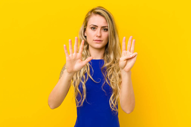 young blonde woman smiling and looking friendly, showing number eight or eighth with hand forward, counting down against yellow wall - Photo, Image