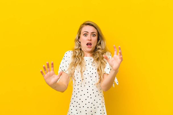 young blonde woman feeling stupefied and scared, fearing something frightening, with hands open up front saying stay away against yellow wall - 写真・画像