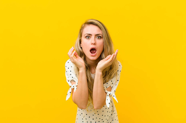 young blonde woman feeling shocked and excited, laughing, amazed and happy because of an unexpected surprise against yellow wall - Foto, Imagem
