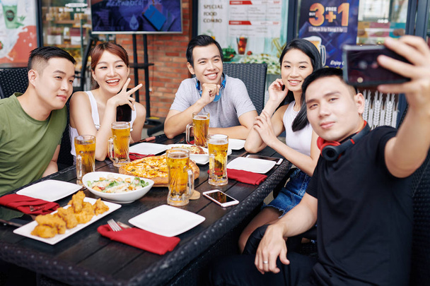 Young Asian man holding mobile phone and making a selfie portrait with his friends during their dinner at the restaurant - Photo, Image