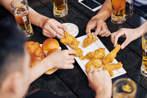 Κοντινό πλάνο των ανθρώπων που κάθονται στο ξύλινο τραπέζι πίνοντας μπύρα και τρώγοντας τηγανητό κοτόπουλο μαζί στο καφέ - Φωτογραφία, εικόνα