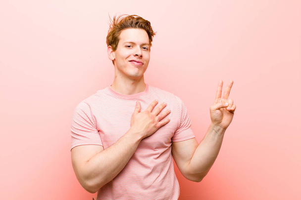 young red head man looking happy, confident and trustworthy, smiling and showing victory sign, with a positive attitude against pink wall - Photo, Image