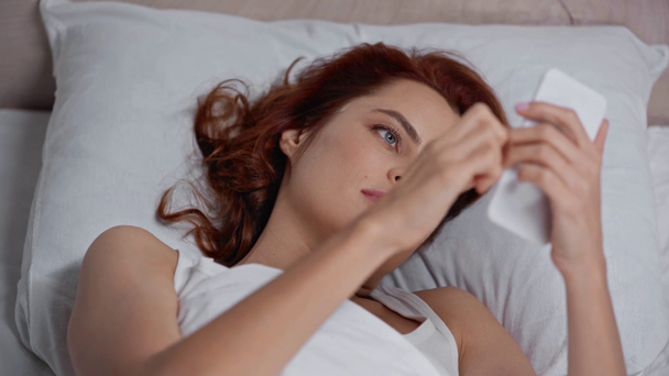 wesoła kobieta na czacie na smartfonie leżąc w łóżku - Materiał filmowy, wideo