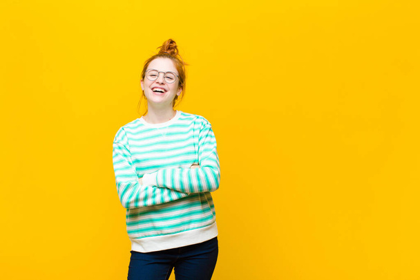 young red head woman looking like a happy, proud and satisfied achiever smiling with arms crossed against orange wall - Foto, immagini