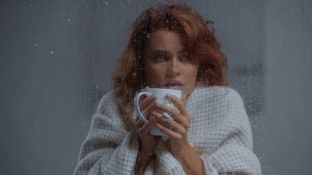 nemocná žena pití teplý nápoj v blízkosti okna skla s kapkami deště - Záběry, video