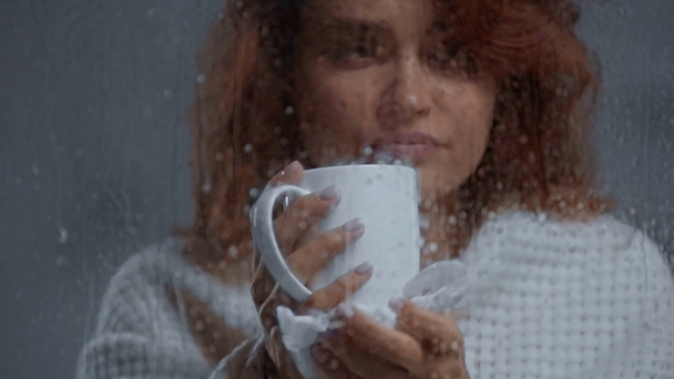 III žena pití ohřívání nápoj v blízkosti okna skla s kapkami deště - Záběry, video
