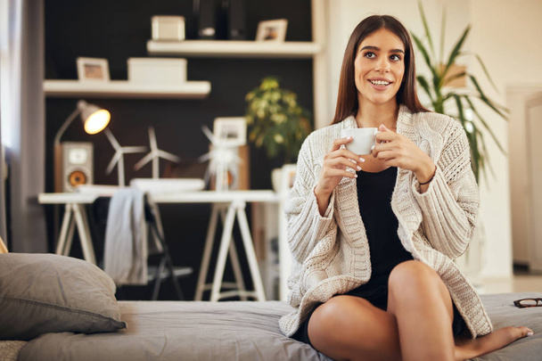Donna caucasica vestita con maglione beige seduta sul letto e con in mano caffè fresco del mattino
. - Foto, immagini