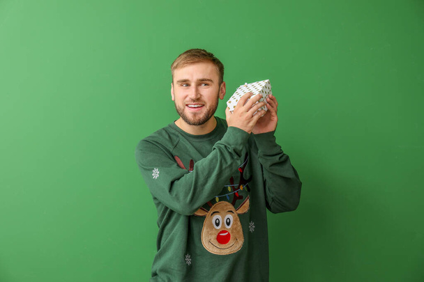 Young man in Christmas sweater and with gift on color background - Photo, Image