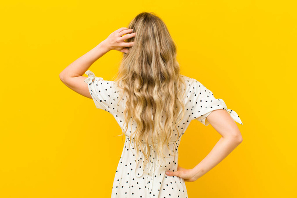 young blonde woman feeling clueless and confused, thinking a solution, with hand on hip and other on head, rear view against yellow wall - Photo, image