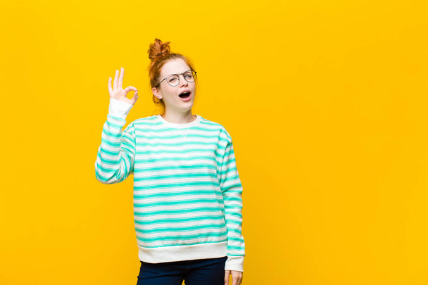 young red head woman feeling successful and satisfied, smiling with mouth wide open, making okay sign with hand against orange wall - Fotó, kép