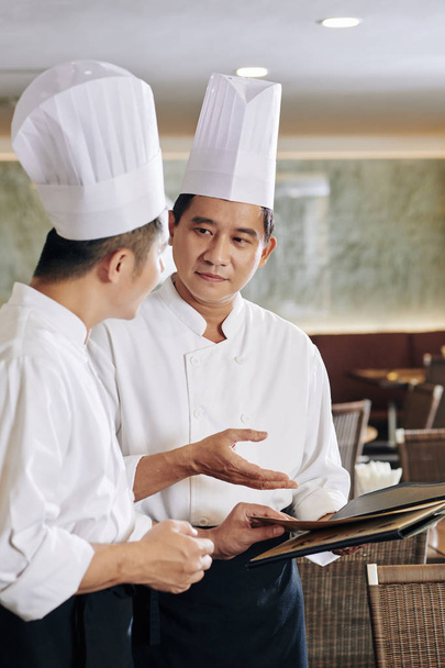 Ασιάτης σεφ με στολή να δείχνει το μενού και να το συζητά μαζί με το συνάδελφό του στο εστιατόριο - Φωτογραφία, εικόνα