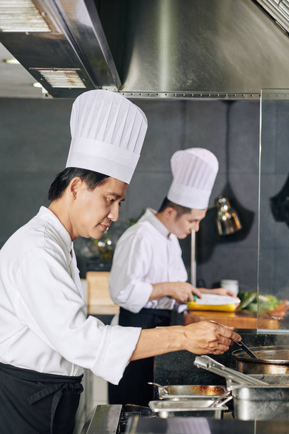 Asiático maduro chef en sombrero blanco de pie cerca de la estufa y la preparación de alimentos junto con su asistente en el fondo en la cocina
 - Foto, Imagen