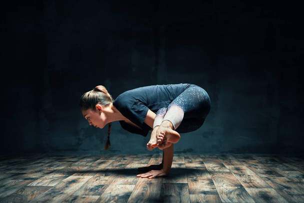 Young  woman practicing yoga doing side crane pose in dark room. Parsva Bakasana exercise.  - Φωτογραφία, εικόνα