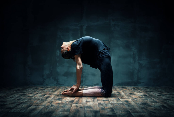 Mladá žena, která cvičí jógu na velbloudech, pózuje v tmavé místnosti. Ustrasana asana. - Fotografie, Obrázek