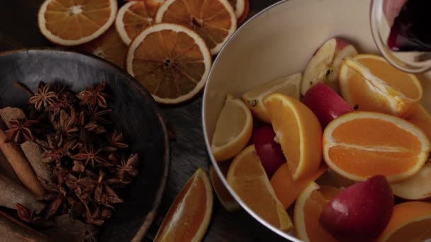 Preparation of mulled wine. Pour red wine in to saucepan on top of lemons, oranges, apples. Anise stars and cinnamon sticks in brown wooden bowl in foreground. Close up top view. - Footage, Video