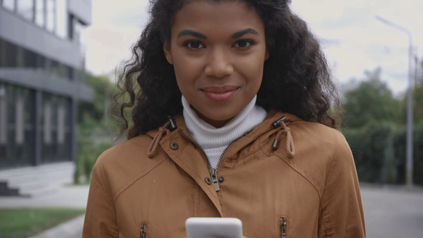 χαμογελαστή Αφροαμερικανή γυναίκα που χρησιμοποιεί smartphone στο δρόμο  - Πλάνα, βίντεο