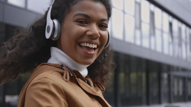 χαμογελαστή Αφροαμερικανή που περπατάει στο δρόμο και ακούει μουσική με ακουστικά - Πλάνα, βίντεο