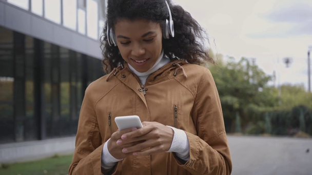 χαμογελαστή Αφροαμερικανή γυναίκα με ακουστικά χρησιμοποιώντας smartphone στο δρόμο  - Πλάνα, βίντεο