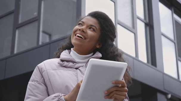 niski kąt widzenia uśmiechnięta Afroamerykanka za pomocą tabletu cyfrowego w pobliżu budynku - Materiał filmowy, wideo
