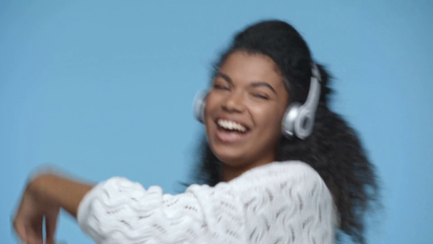 happy african american woman listening music in headphones and dancing isolated on blue - Materiaali, video