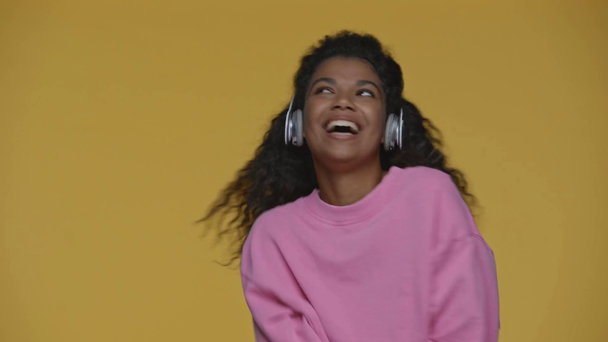 happy african american girl listening music in headphones, dancing with smartphone isolated on yellow - Footage, Video