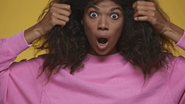 wstrząśnięta afrykańska dziewczyna w różowej bluzie chowająca się za włosami odizolowanymi na żółto - Materiał filmowy, wideo