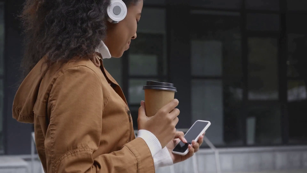щаслива афроамериканка в навушниках, що йдуть і п'ють з паперової чашки, використовуючи смартфон на вулиці
 - Кадри, відео