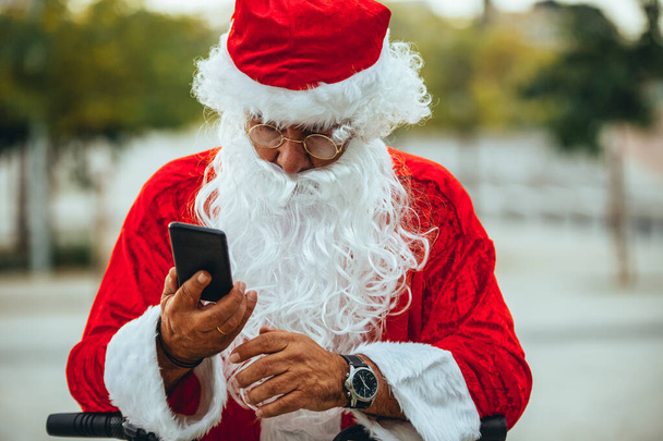 Foto stock di Babbo Natale che parla con un telefono cellulare con un parco dietro sfocato. Tempo di Natale
 - Foto, immagini
