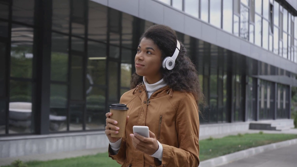 щаслива афроамериканка в навушниках, що йдуть і п'ють з паперової чашки, використовуючи смартфон на вулиці
 - Кадри, відео