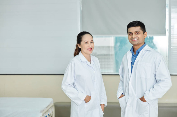 病院に立ちながらカメラで微笑む白衣姿のインド人男性医師とアジア人女性医師の肖像 - 写真・画像