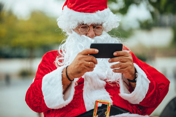 Foto de Santa Claus conversando com um telefone celular com um parque atrás desfocado. Tempo de Natal
 - Foto, Imagem