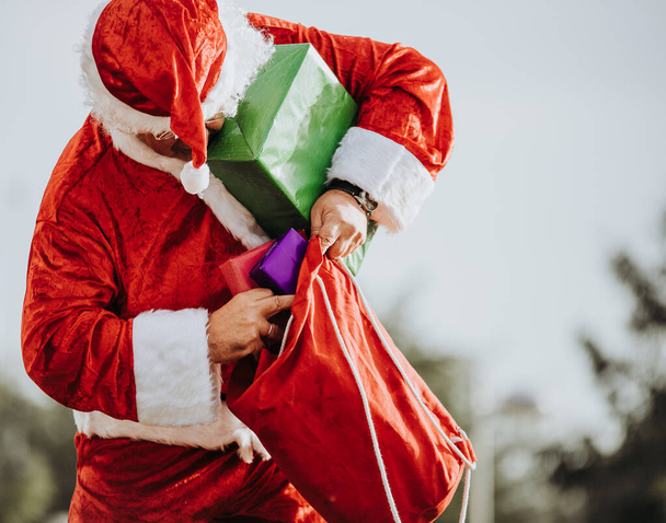 Άγιος Βασίλης χωρίς γενειάδα στέκεται βάζοντας δώρα σε μια κόκκινη τσάντα - Φωτογραφία, εικόνα