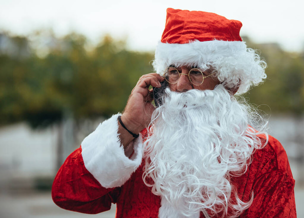 Foto en stock de Santa Claus hablando con un teléfono móvil con un parque detrás desenfocado. Tiempo de Navidad
 - Foto, imagen