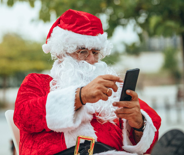 サンタクラスのストックフォトは、公園の後ろに焦点を当てていない携帯電話と話しています。クリスマスタイム - 写真・画像