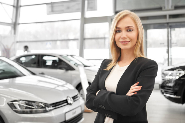 Female car dealer with folded arms standing in auto salon - 写真・画像