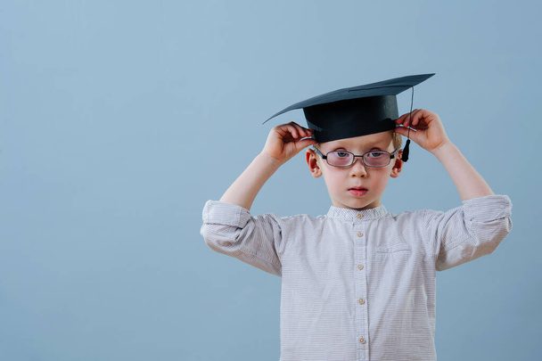 Redhead first grader boy in glasses fixing his student hat over blue background - Photo, image