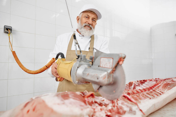 Butcher using electric knife for cutting pork at factory. - Zdjęcie, obraz