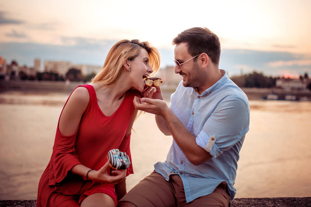 Szczęśliwa młoda para bawiąca się na świeżym powietrzu, jedząc pączki. - Zdjęcie, obraz