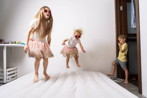Little girls in sunglasses and identical dresses are jumping on a white mattress. - Photo, Image