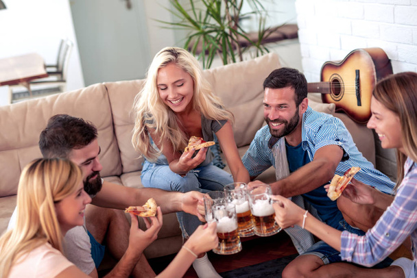 Φίλοι διασκεδάζουν μαζί στο σπίτι, τρώνε πίτσα και πίνουν μπύρες. - Φωτογραφία, εικόνα