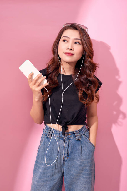 Мода довольно крутая улыбающаяся девушка, расслабляющаяся, слушающая музыку в наушниках на розовом фоне. - Фото, изображение